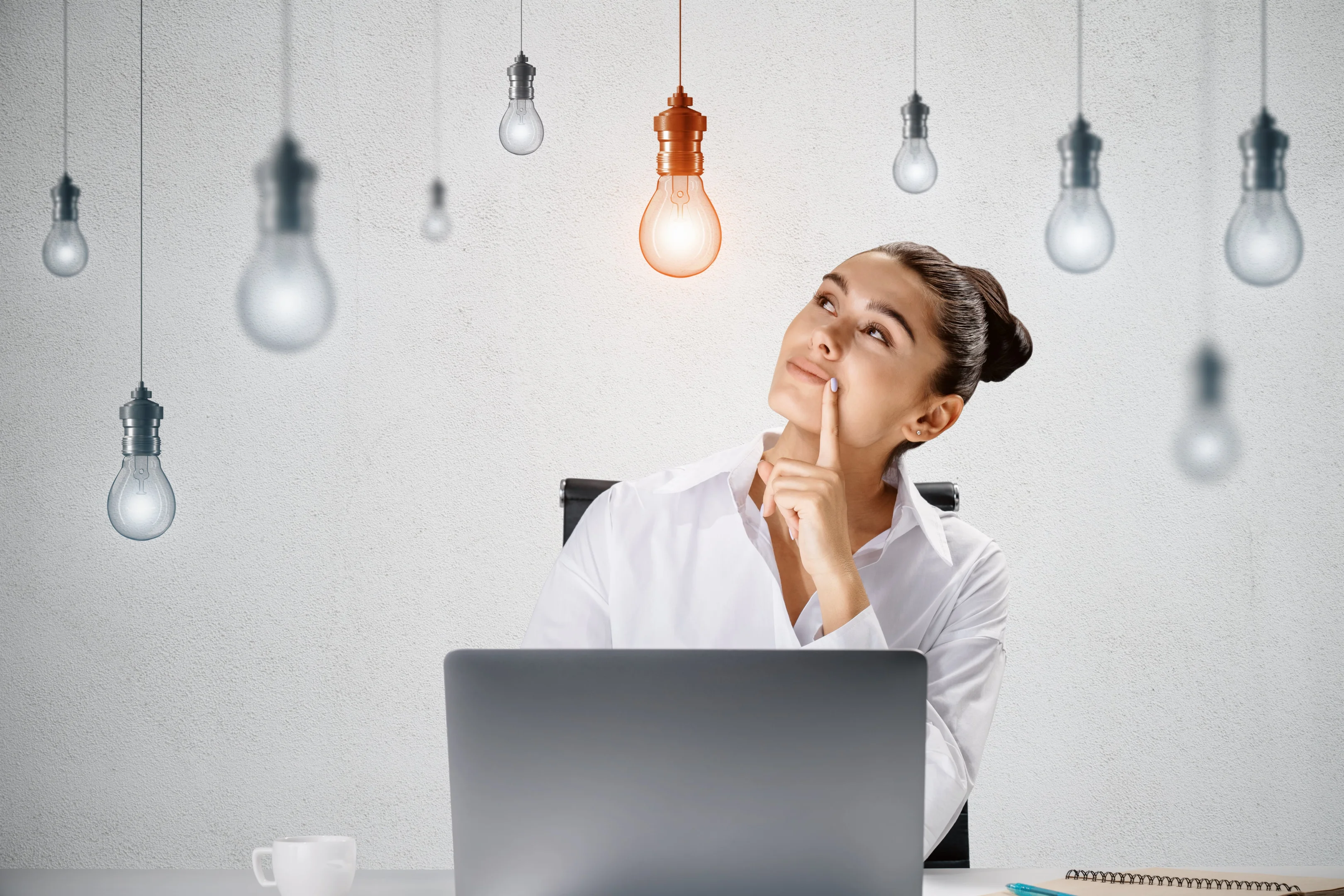 Photo creative idea and inspiration concept with pensive young woman sitting in front of laptop at light wall background with hanging from the top switched on yellow lightbulb among white lightbulbs